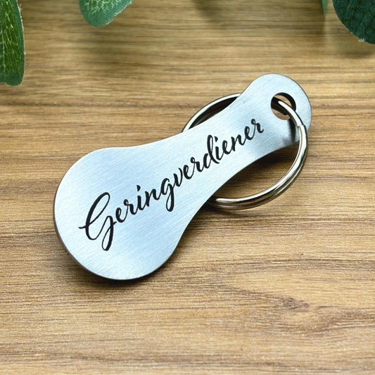 Lustiger Einkaufswagenchip aus Edelstahl mit Gravur „Geringverdiener“ – witziger Schlüsselanhänger auf Holzoberfläche