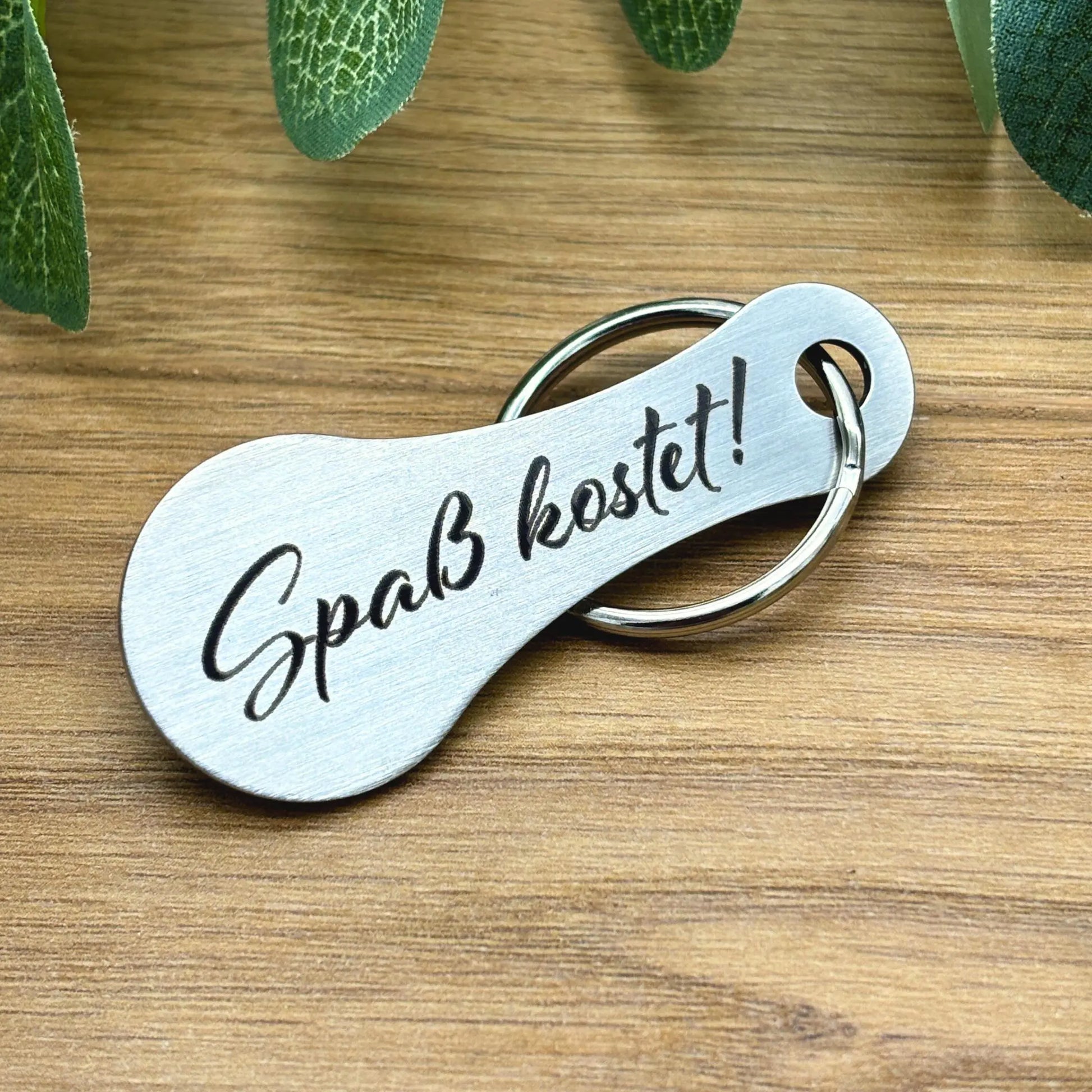 Einkaufswagenlöser „Spaß kostet!“ aus Edelstahl mit Gravur auf Holztisch