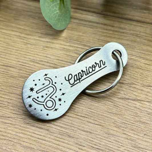 Einkaufswagenlöser Schlüsselanhänger aus Metall mit Gravur des Sternzeichens Capricorn (Steinbock) auf Holzuntergrund – elegantes Geschenk für Astrologie-Fans.